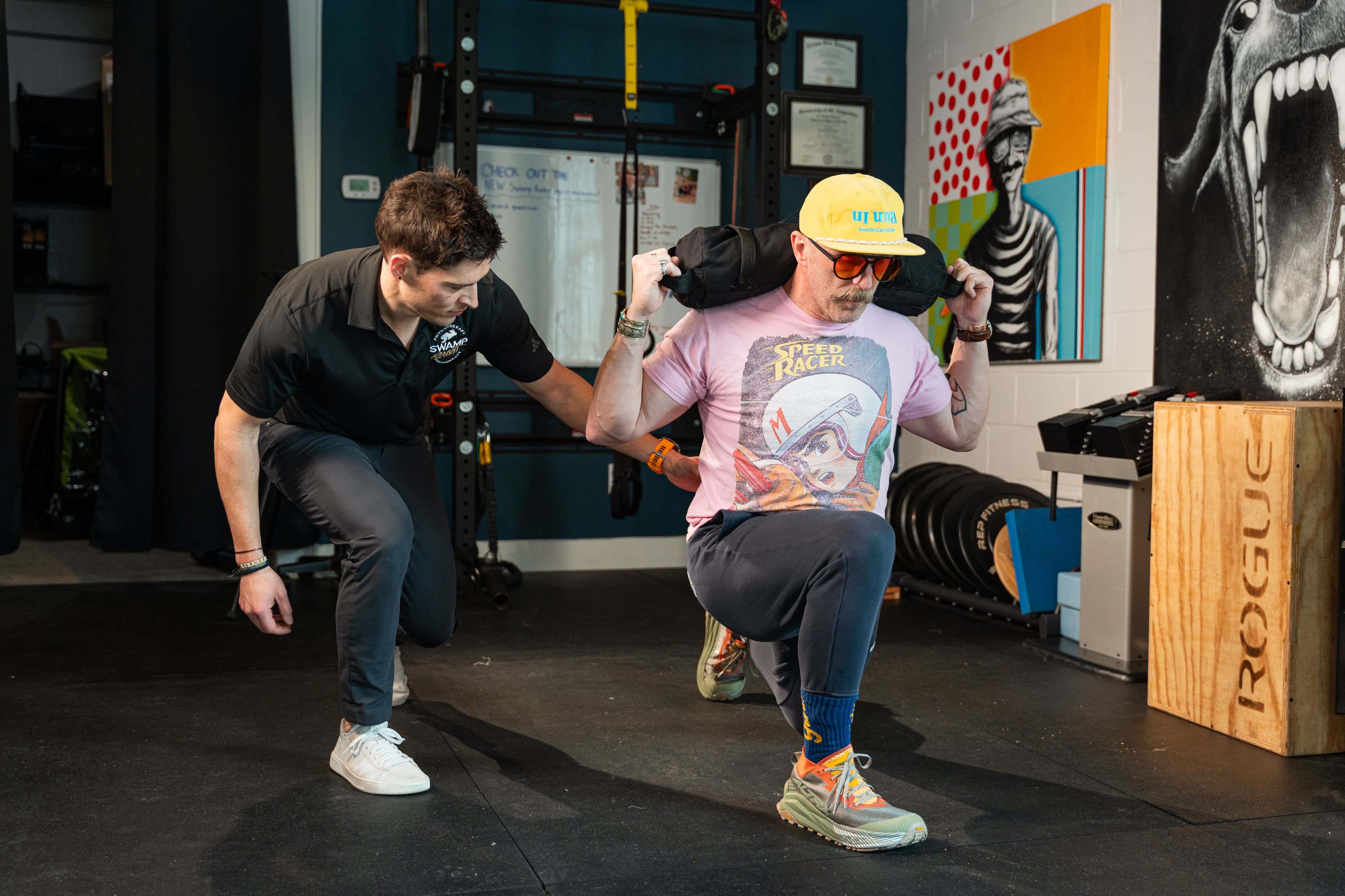 Physical therapist coaching a patient through a weighted lunge exercise with a sandbag in the gym at Swamp Rabbit Physiotherapy in Greenville, SC
